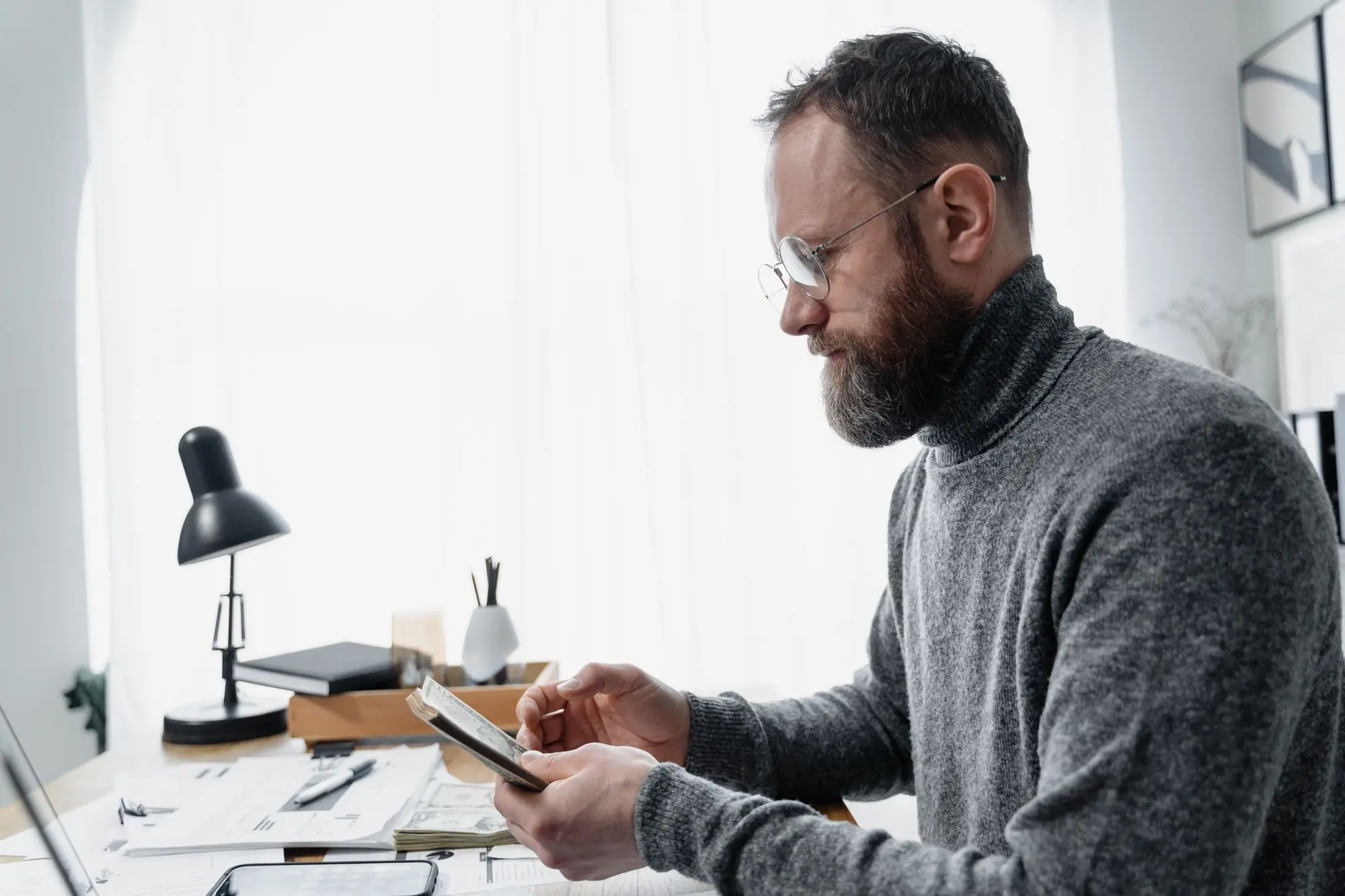 Business owner at his home office desk checking figures on his phone beside a pile of settlement and business papers