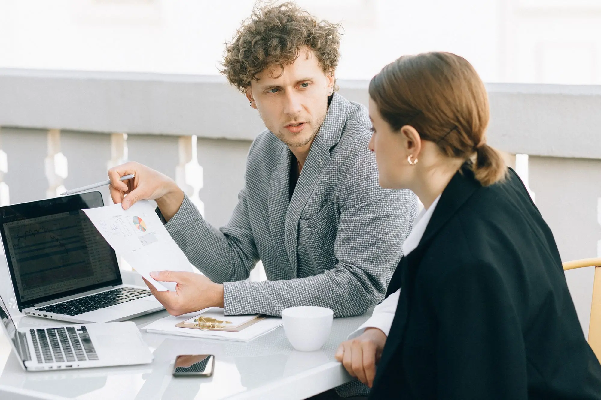 Two business partners at an outdoor table going over printed figures and a laptop while deciding whose property should back a loan